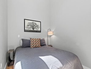 A bedroom with a bed, a bedside table, a lamp, a wall-mounted picture, and a cat bed. - Hot Tub+Fire Pit, On the River, WiFi, Grill, Cozy! (Burnsville)