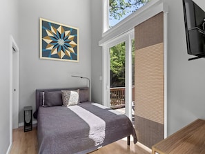 A bedroom with a bed, a wall-mounted TV, a window with a view of greenery, and a door leading to an outdoor area.