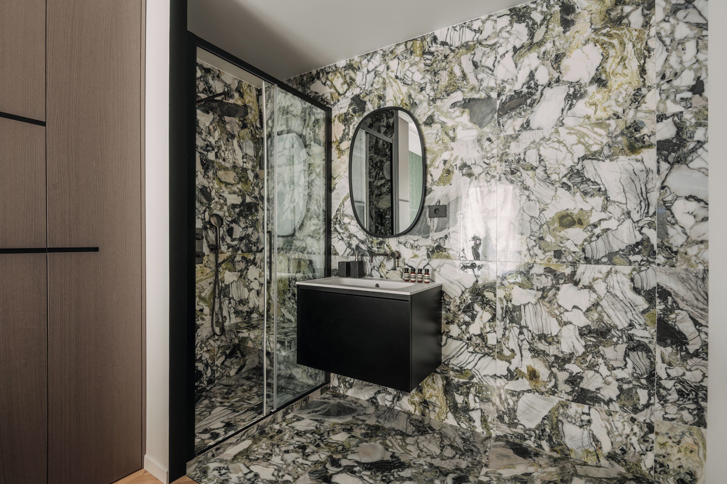 A bathroom with a marble wall, a black vanity, and a glass shower enclosure.