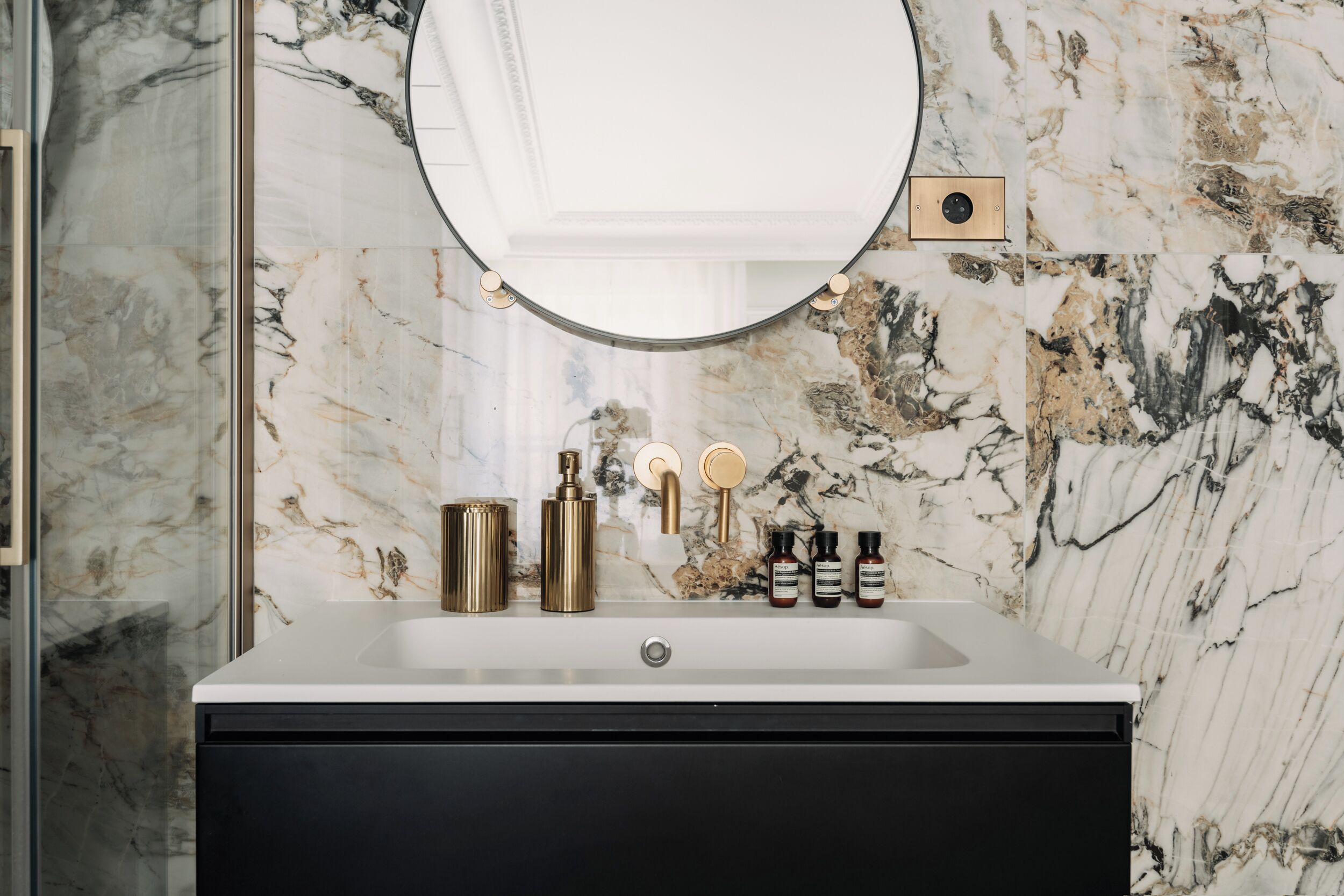 Un baño con salpicadero de mármol, un espejo redondo, un mueble de tocador negro y un lavabo con detalles dorados.