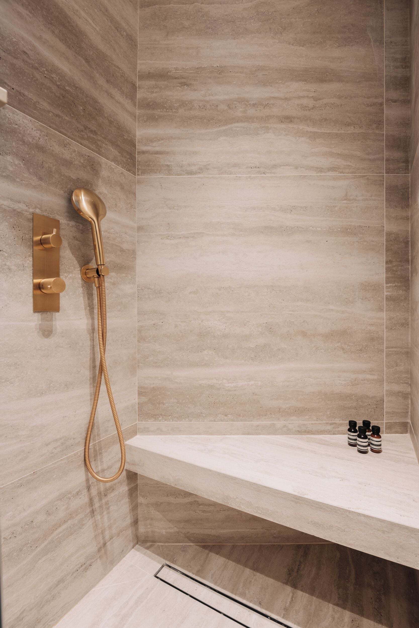 A modern shower with a gold showerhead and control panel, a built-in bench, and a small shelf with bottles.