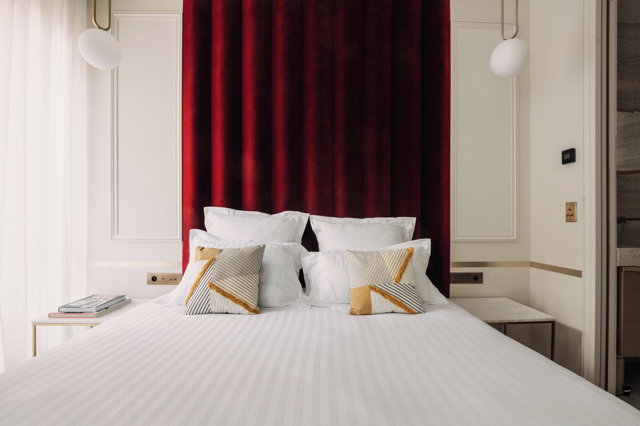 A neatly made bed with white linens and decorative pillows, a bedside table with a book, and a red curtain.