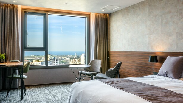 Une chambre d’hôtel avec une grande fenêtre, un lit, un bureau et une chaise.