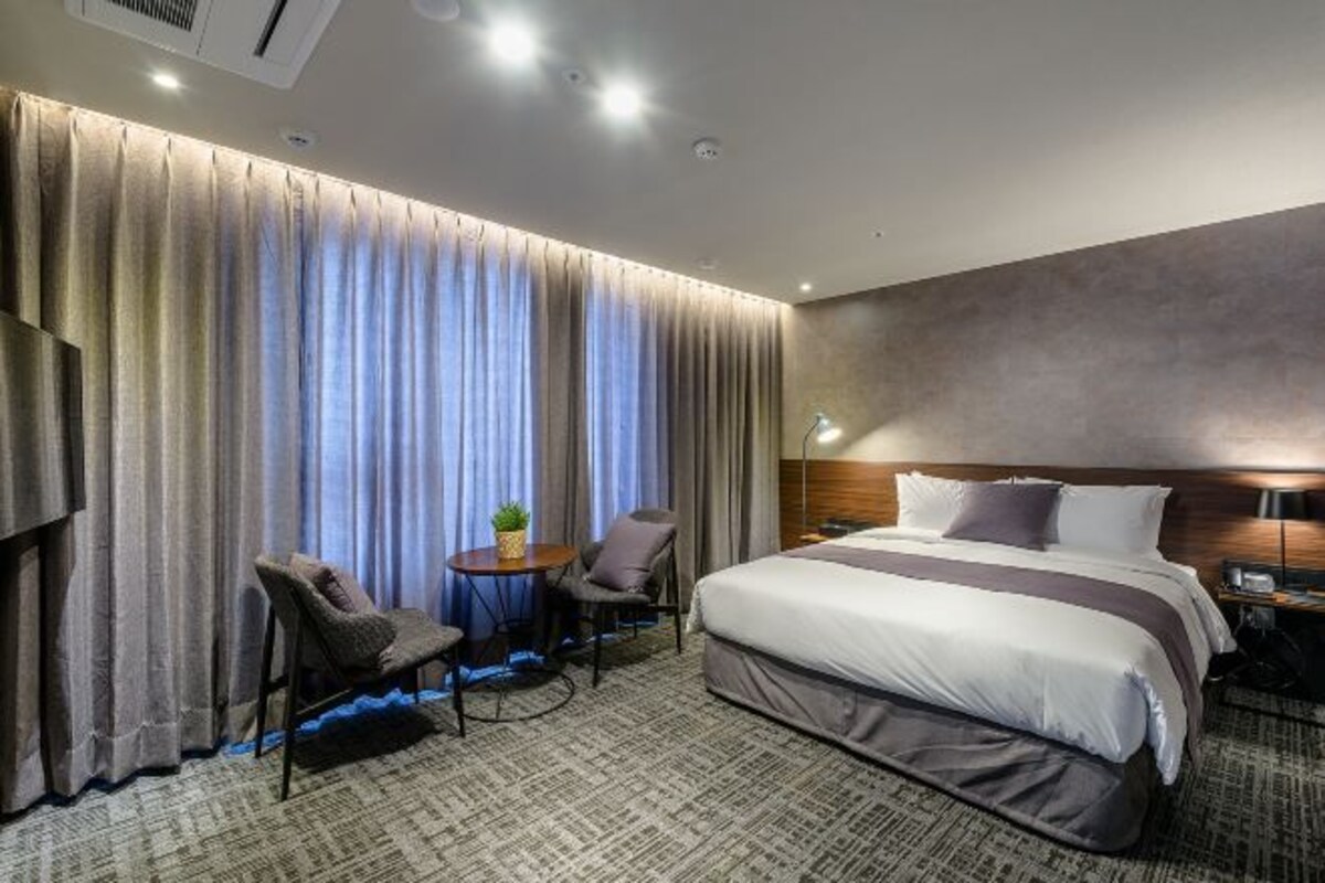 A hotel room with a large bed, a desk with a chair, a small table with a plant, and a large window with curtains.