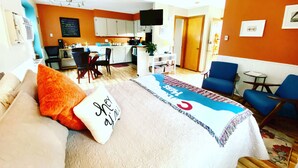 Ett hotellrum med en säng, ett skrivbord, en stol och ett litet kök i bakgrunden.