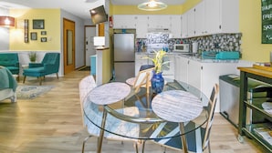 Cocina moderna con armarios blancos, refrigerador de acero inoxidable y microondas. Hay una zona de comedor con mesa de vidrio y sillas, y una planta pequeña sobre la mesa.