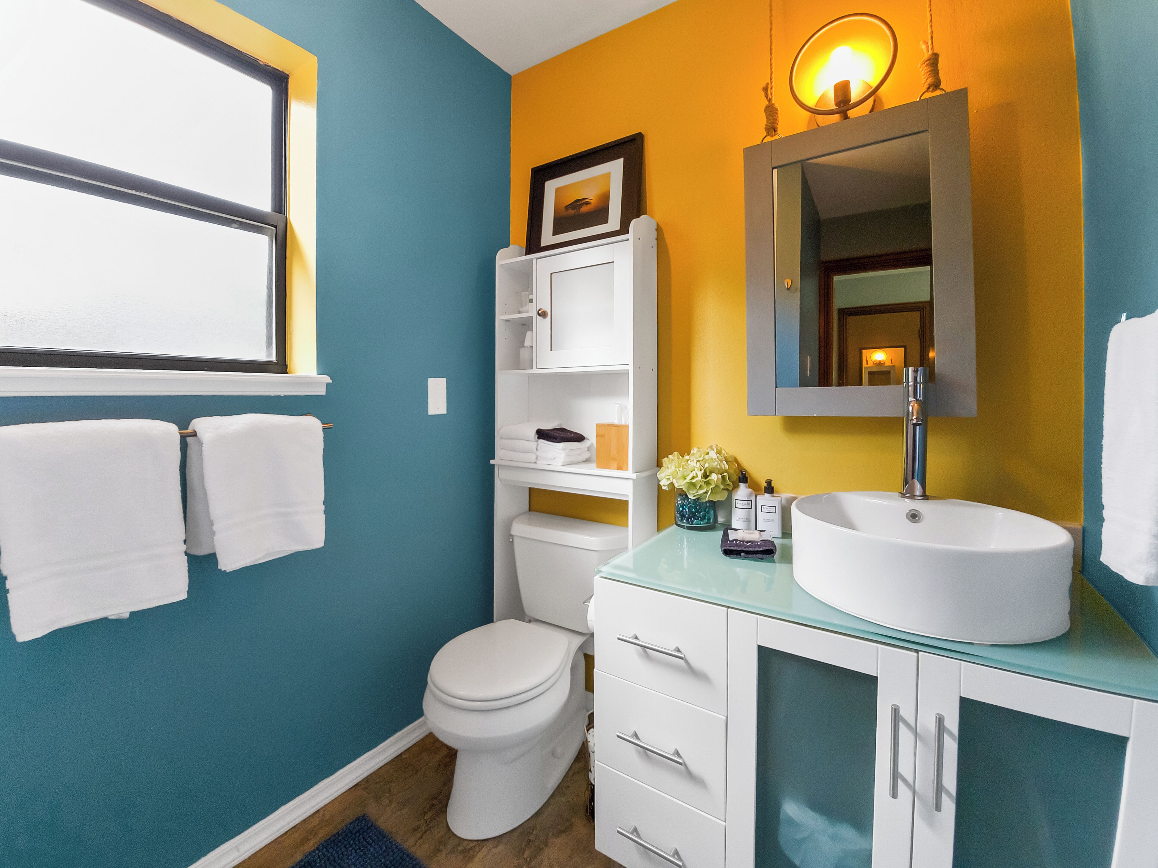 A bathroom with a toilet, a sink, and a mirror. There are towels hanging on the wall and a window above the toilet.