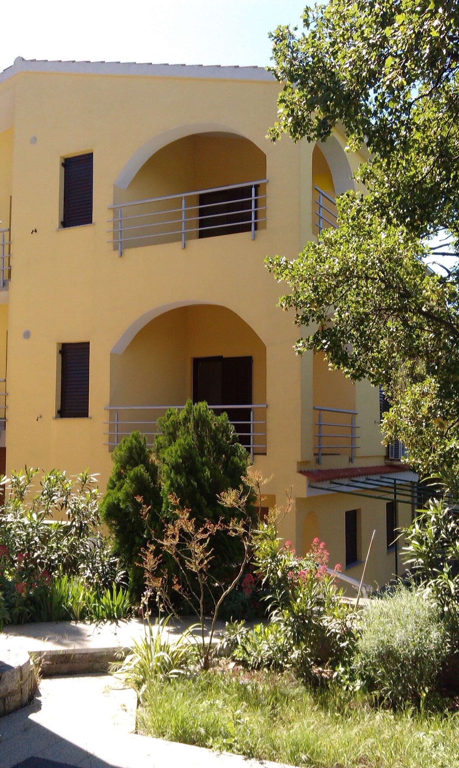 A yellow building with three balconies, surrounded by greenery and a pathway.