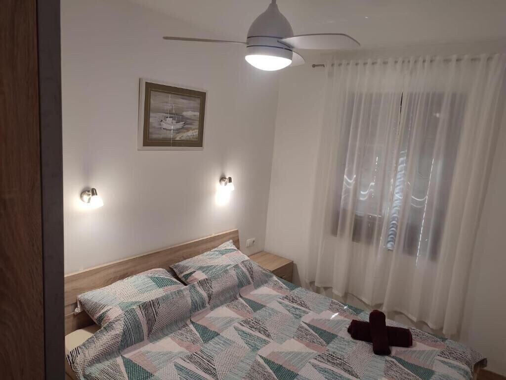 Une chambre à coucher comprenant un lit, un ventilateur de plafond, une table de chevet, une lampe fixée au mur et une fenêtre avec des rideaux légers.