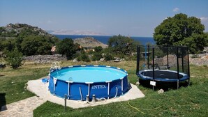 Una piscina fuori terra blu con un trampolino nelle vicinanze, immersa in un’area erbosa con vista panoramica sul mare e sulle colline sullo sfondo.