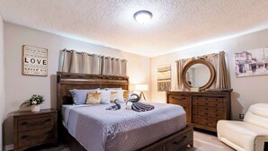 Un dormitorio con cama de madera, mesita de noche, cómoda, silla y pared con una cita enmarcada.