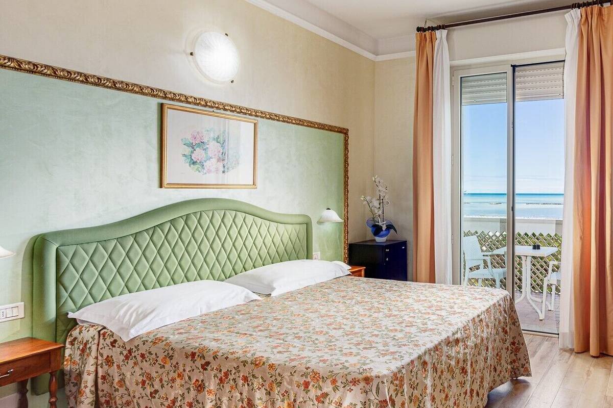 Quarto de hotel com cabeceira verde, colcha florida, mesa de cabeceira, abajur, quadro emoldurado na parede e vista para o mar por uma porta de correr.