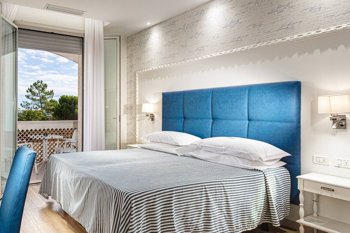 Quarto de hotel com uma cama grande, varanda com vista e uma mesa de cabeceira branca.