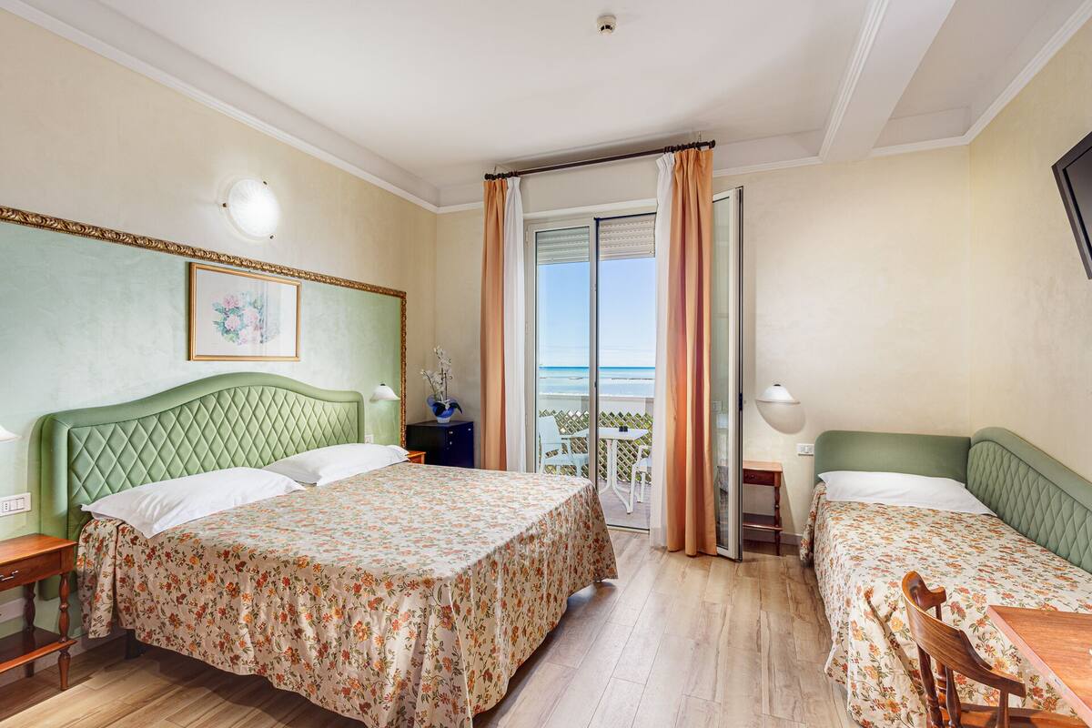 Quarto de hotel com duas camas, uma varanda e vista para o mar, e uma pequena mesa com uma cadeira.