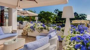 Un área de estar al aire libre con mobiliario de mimbre blanco, incluyendo sofás y una mesa de centro de vidrio. Hay sombrillas que proporcionan sombra, y se ve un letrero del Hotel International al fondo.
