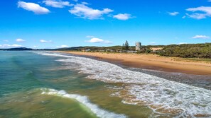 Une plage côtière avec un ciel dégagé, un rivage sablonneux et quelques bâtiments visibles au loin.