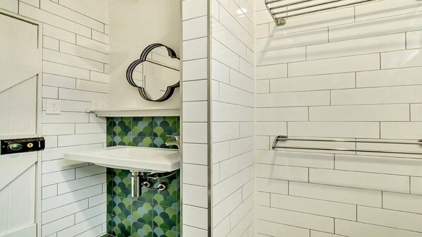 Un baño con azulejos blancos tipo metro, un salpicadero de azulejos estampados verdes, un lavamanos blanco, un espejo y un perchero.