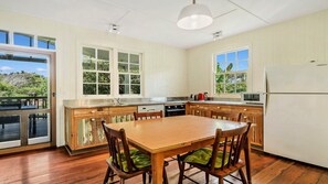A kitchen with a wooden table and chairs, a refrigerator, and a window with a view of greenery. - Moomba On Main - Beachfront cottage in Wooli (WOOLI)