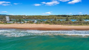 A coastal landscape with a sandy beach, ocean waves, and a distant view of a lake surrounded by greenery and buildings.