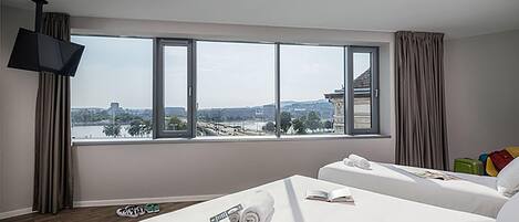 A hotel room with a large window, a flat-screen TV, a bed, a chair, and a table with a towel and book.