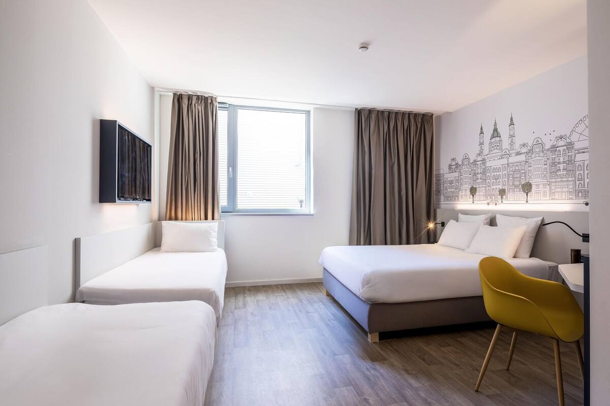 A hotel room with two single beds, a desk, a chair, a window with curtains, and a wall mural.