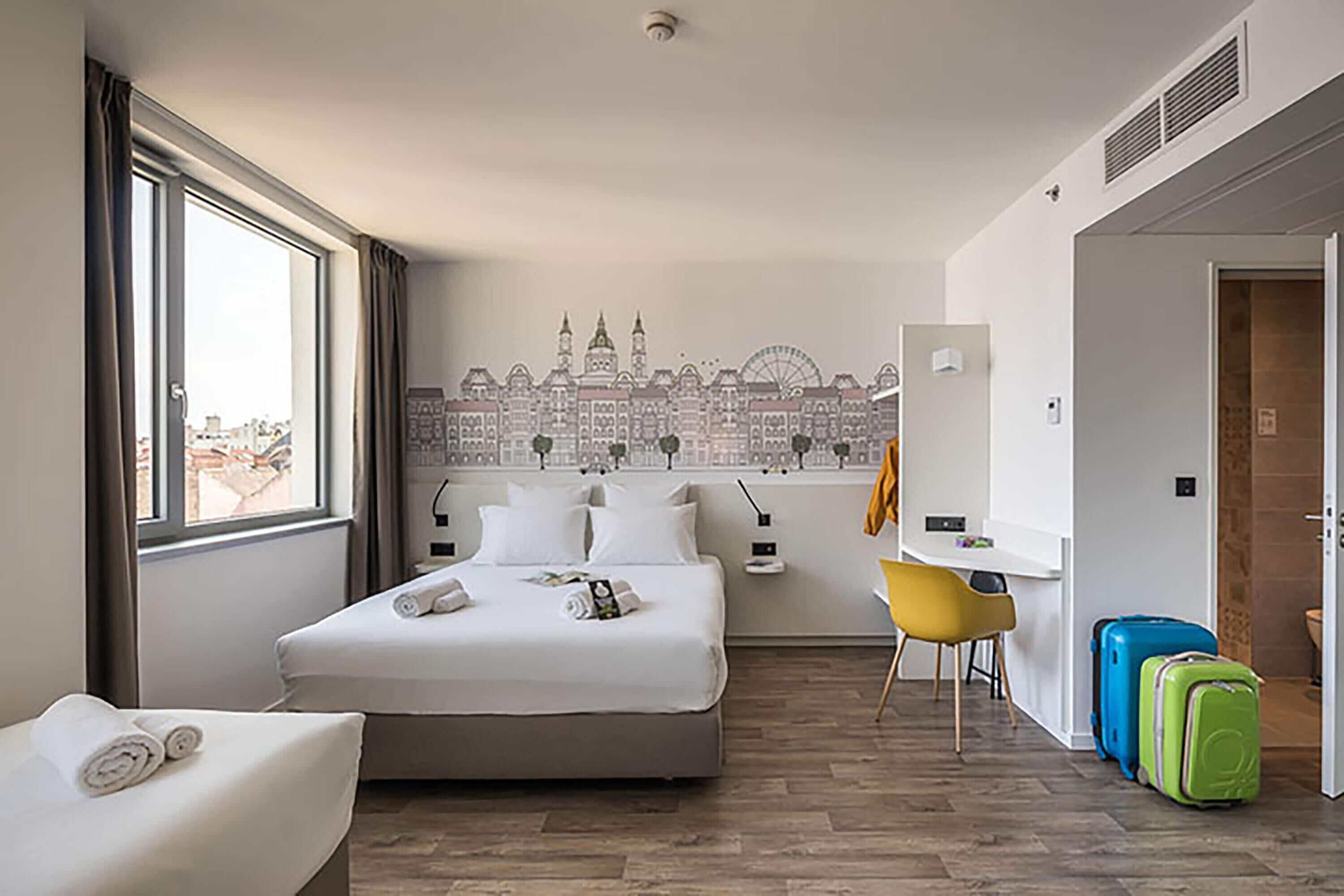 Quarto de hotel com uma cama, uma escrivaninha, uma cadeira e duas malas.