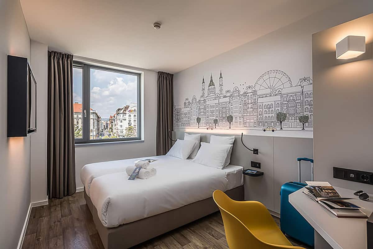 A hotel room with a bed, a desk with a chair, a window with curtains, a wall mural, and a cityscape view.
