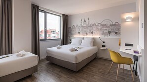 Habitación de hotel con una cama grande, un escritorio, una silla y un mural de un paisaje urbano en la pared.