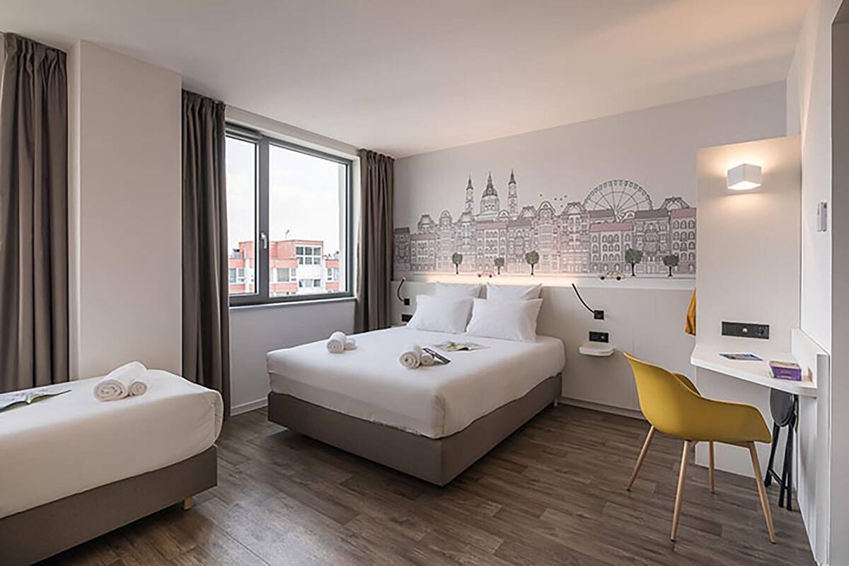 A hotel room with a large bed, a desk, a chair, and a cityscape mural on the wall.