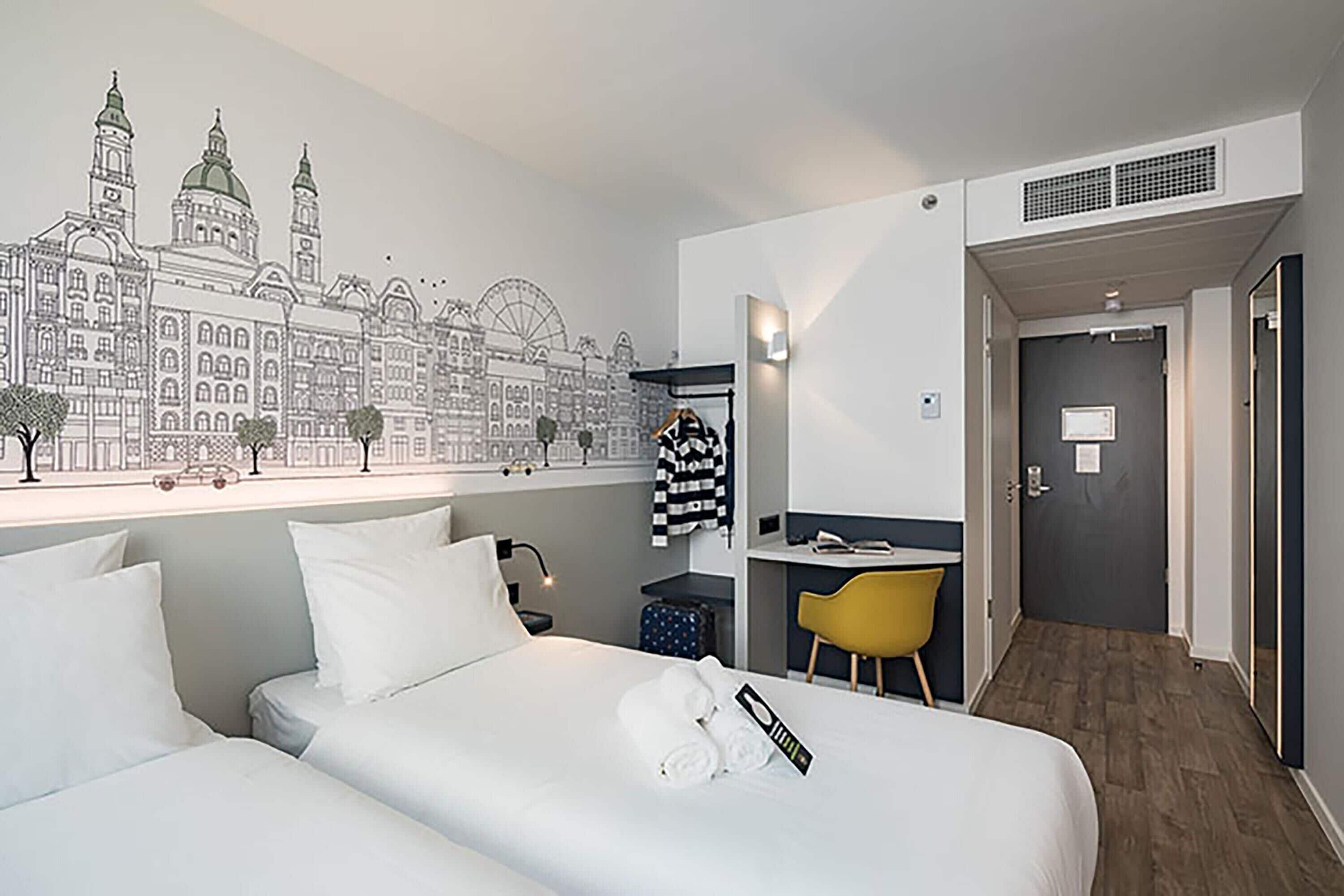 A hotel room with two beds, a desk, a chair, and a mural of a cityscape.