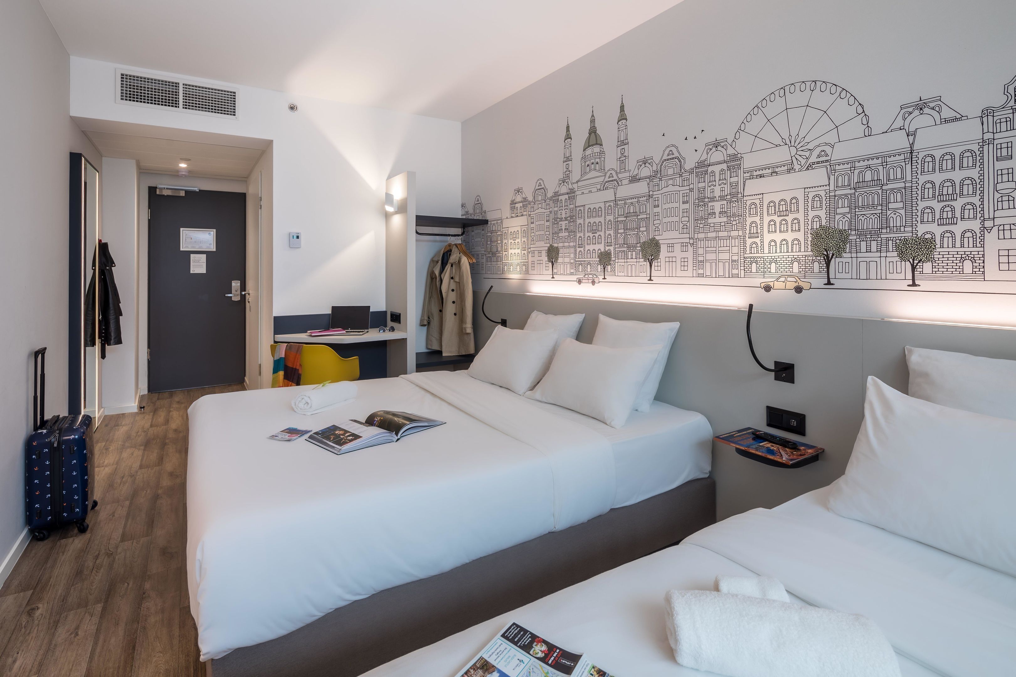 A hotel room with two beds, a cityscape mural, a desk with a chair, and a coat rack.