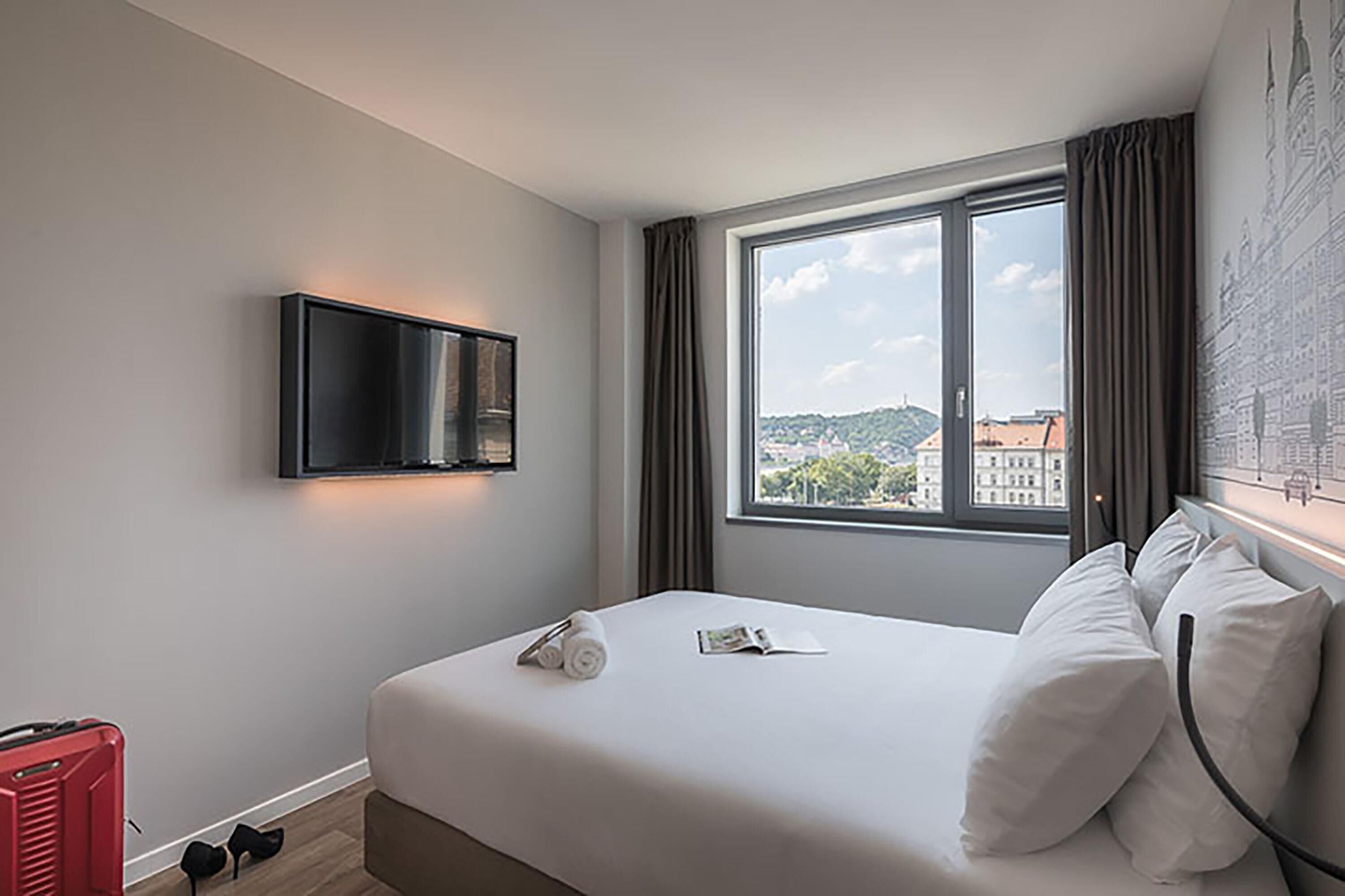 Ein Hotelzimmer mit Bett, Flachbildfernseher an der Wand, einem Fenster mit Vorhängen und einem roten Koffer auf dem Boden.