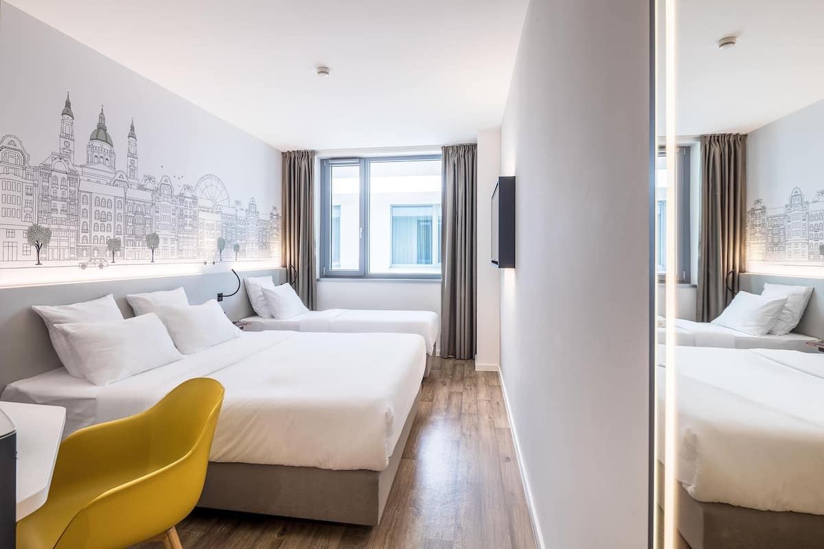 A hotel room with two beds, a yellow chair, a cityscape mural, and a large window with curtains.