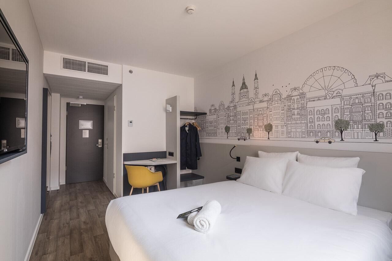 Ein Hotelzimmer mit Bett, Schreibtisch, Sessel und einer Wandmalerei einer Stadtansicht.
