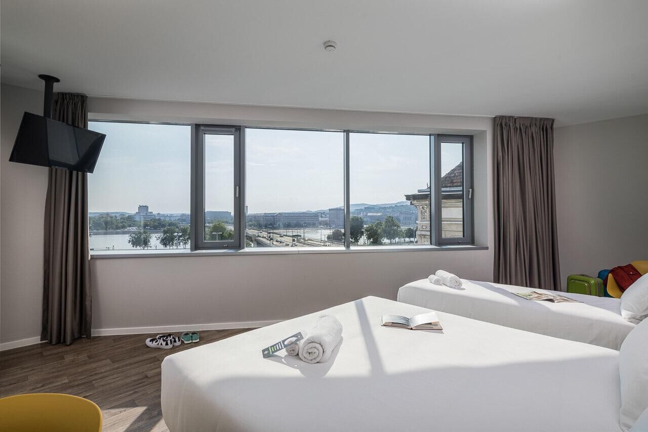 A hotel room with two beds, a TV, and a large window with a city view.