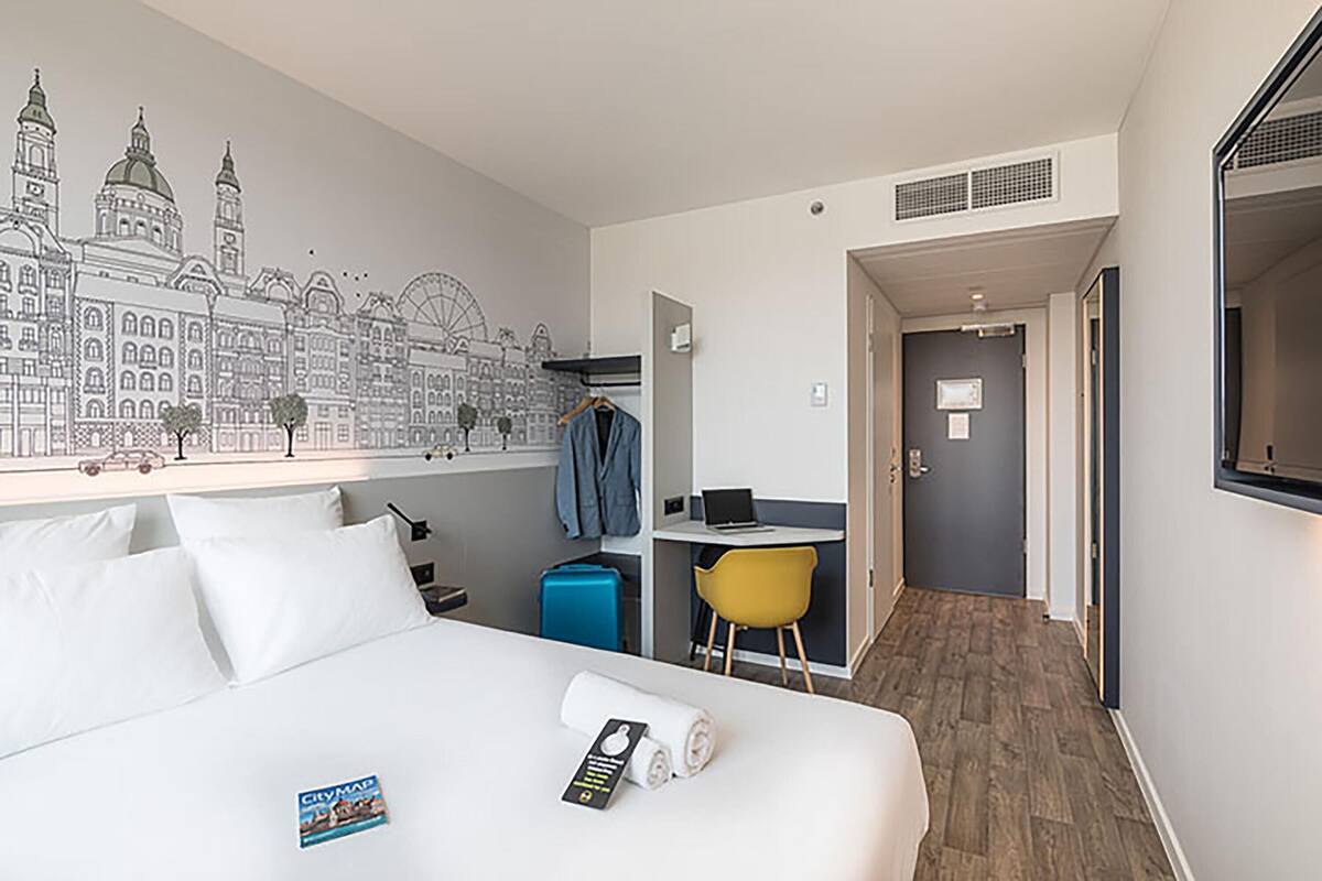 A hotel room with a bed, a desk, a chair, a suitcase, and a wall mural.