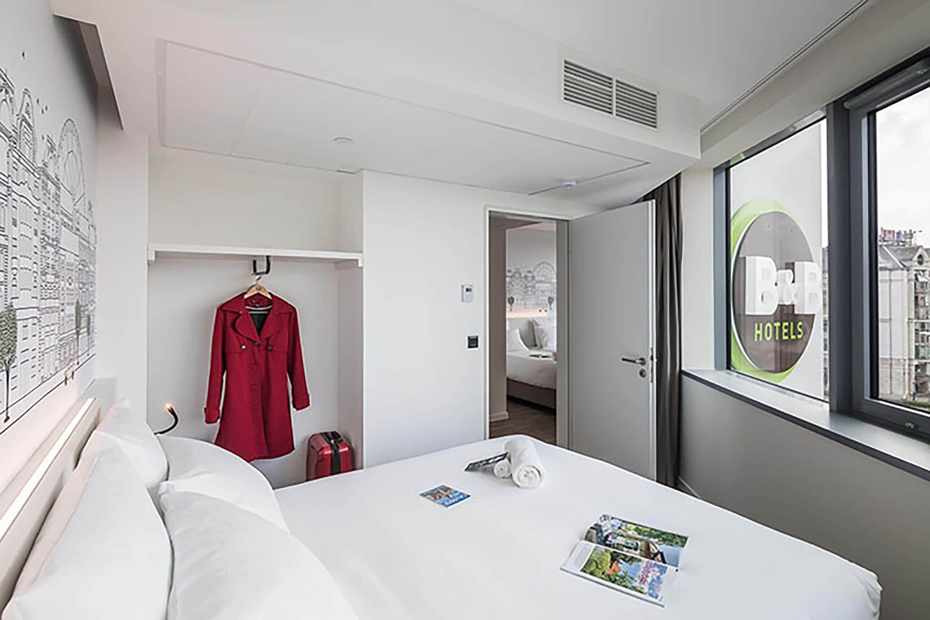 Habitación de hotel con cama, un abrigo rojo colgado en la pared, una maleta y una ventana con vistas a edificios.