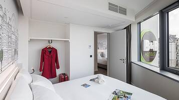 Habitación de hotel con cama, un abrigo rojo colgado en la pared, una maleta y una ventana con vistas a edificios.