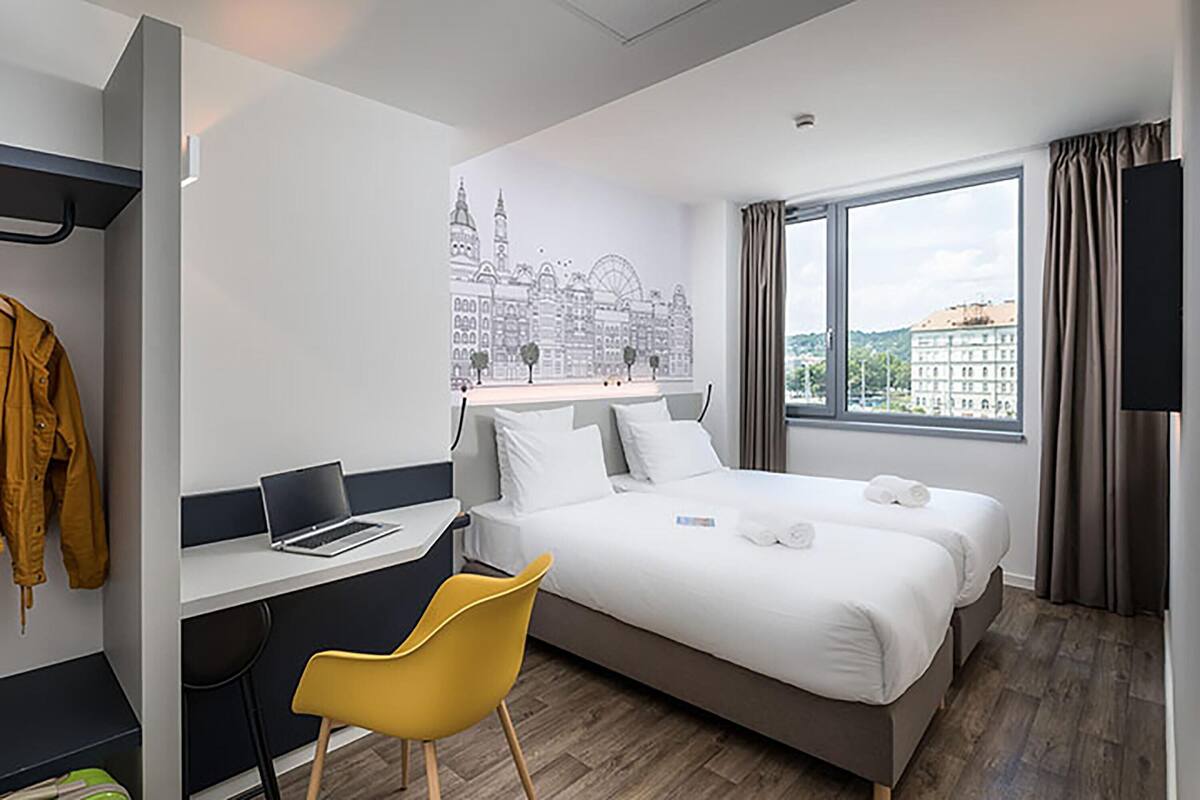 A hotel room with a bed, a desk with a laptop, a chair, and a cityscape mural on the wall.
