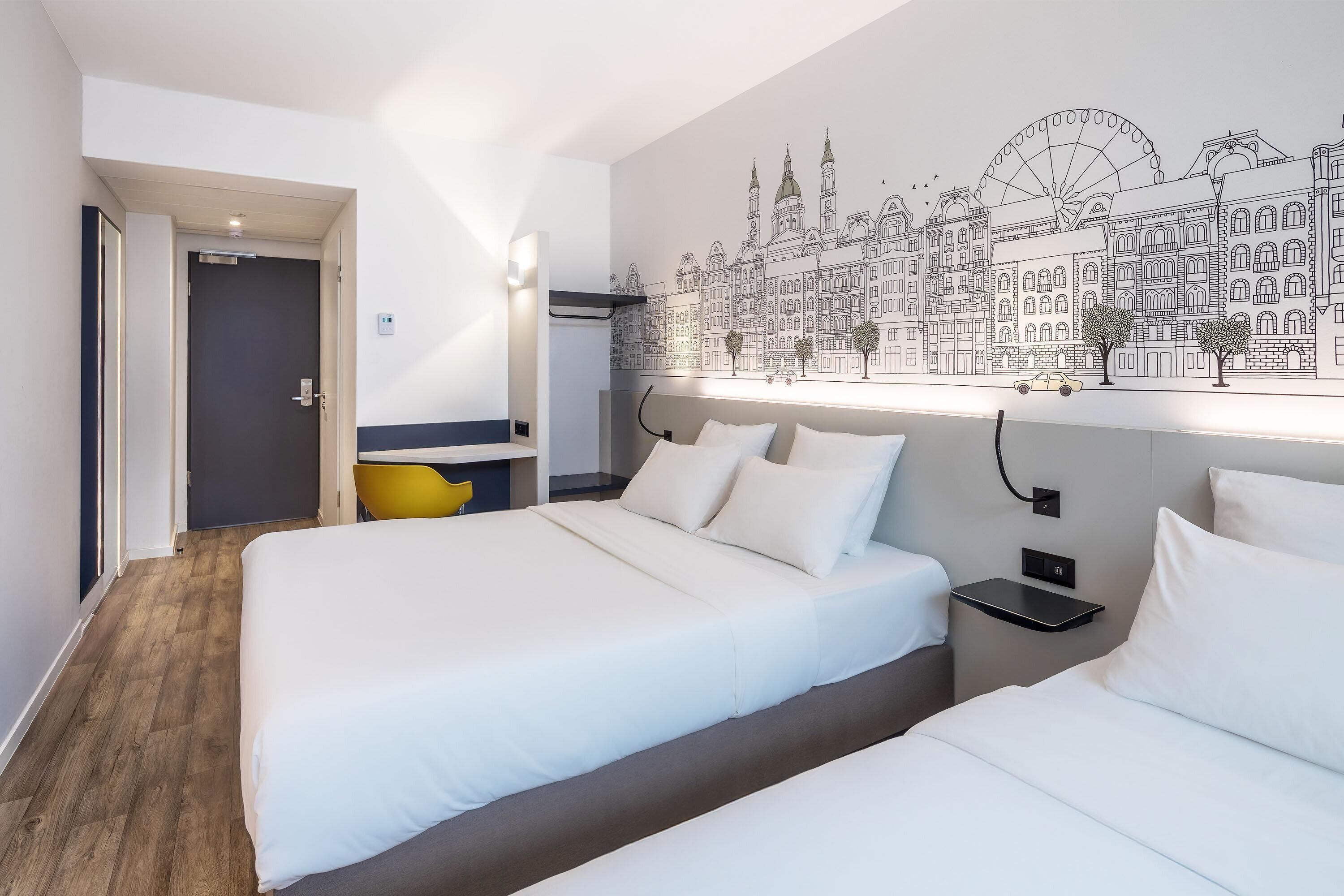 A hotel room with two beds, a cityscape mural, a wooden floor, and a small table with a chair.