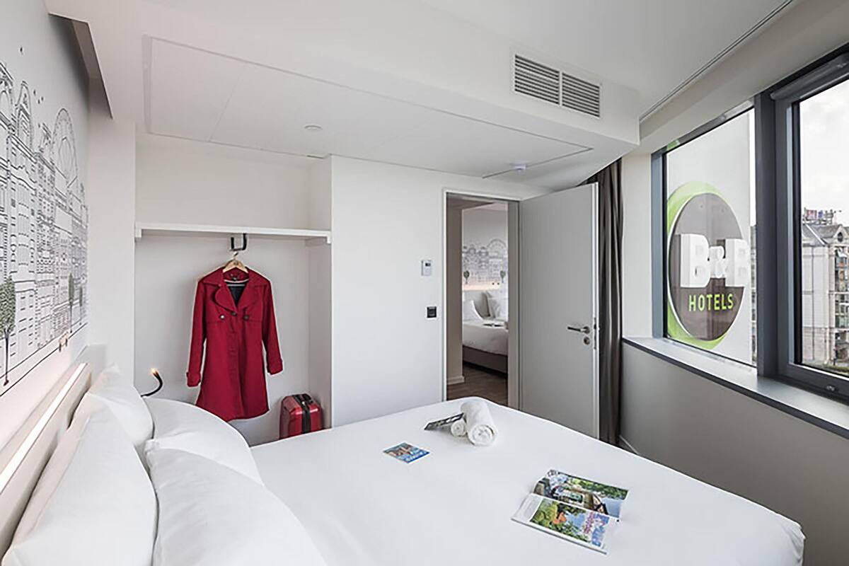 A hotel room with a bed, a red coat on a hanger, a suitcase, and a window with a view of buildings.