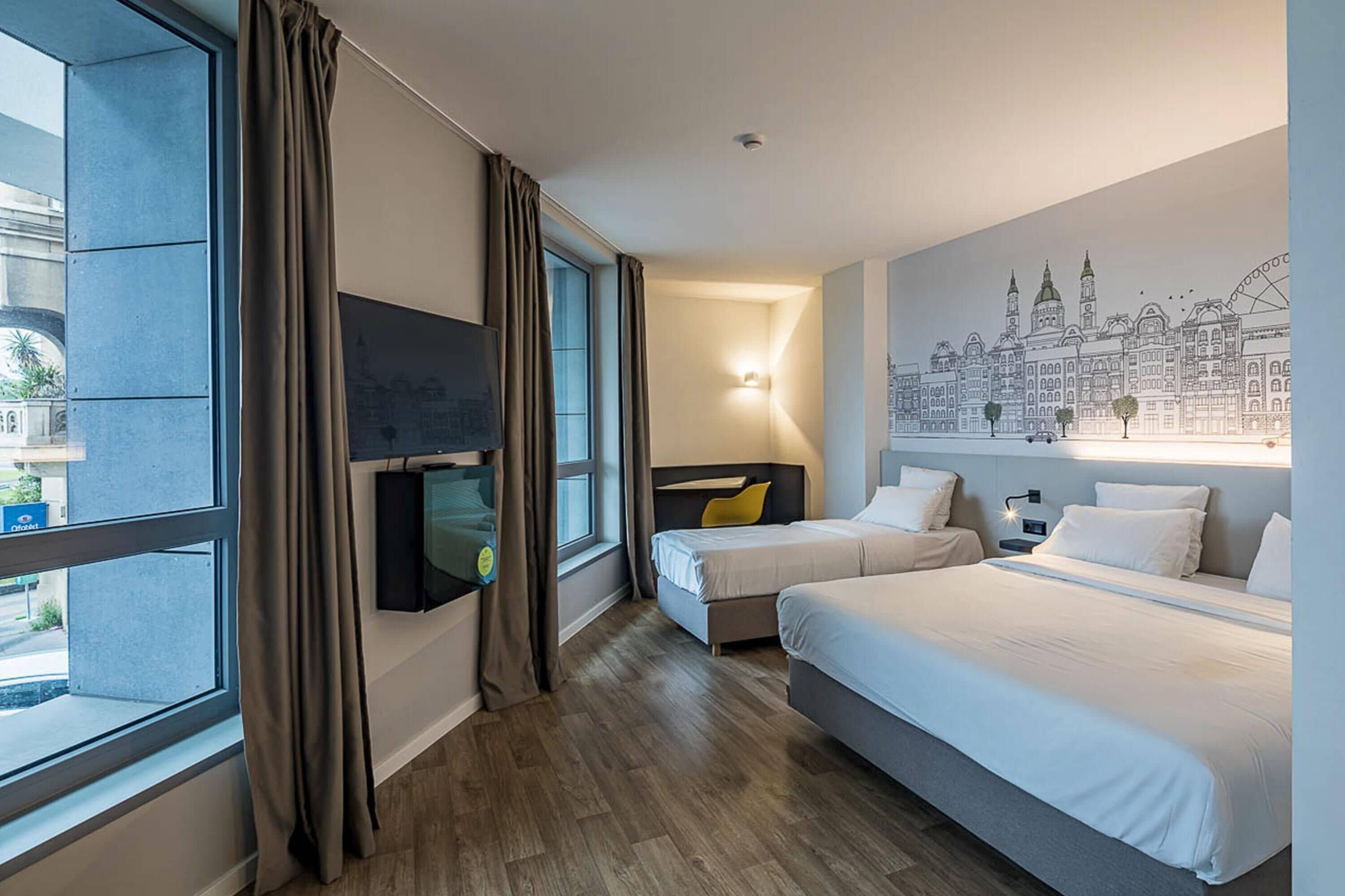 Une chambre d’hôtel avec deux lits, une télévision, une fresque représentant un paysage urbain et une fenêtre avec des rideaux.