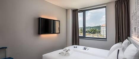 A hotel room with a bed, a flat-screen TV mounted on the wall, a window with curtains, and a view of buildings outside.