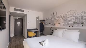 Habitación de hotel con cama, escritorio, silla y un mural en la pared que representa un paisaje urbano.