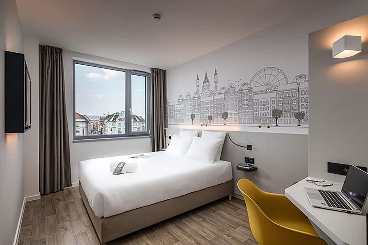 A hotel room with a bed, a desk with a laptop, a chair, and a cityscape mural on the wall.