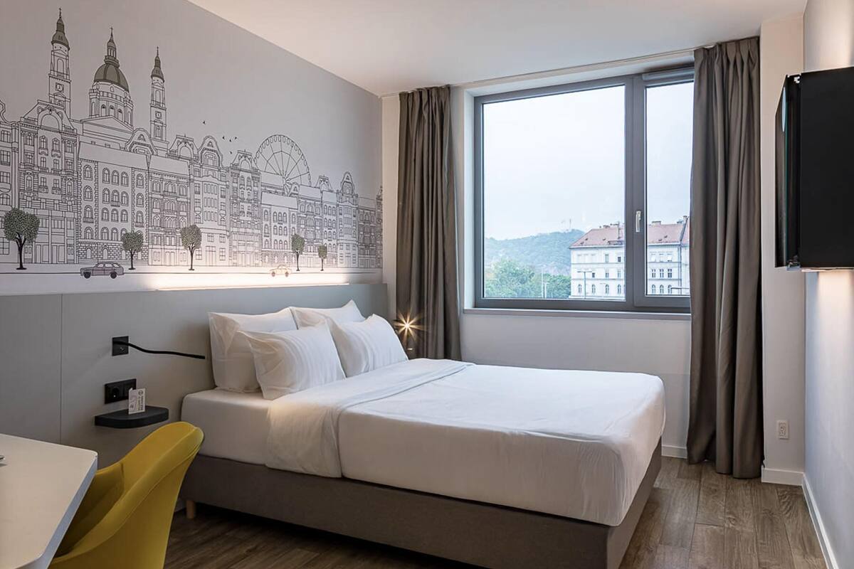 A hotel room with a bed, a desk, a chair, a wall mural, and a window with curtains.