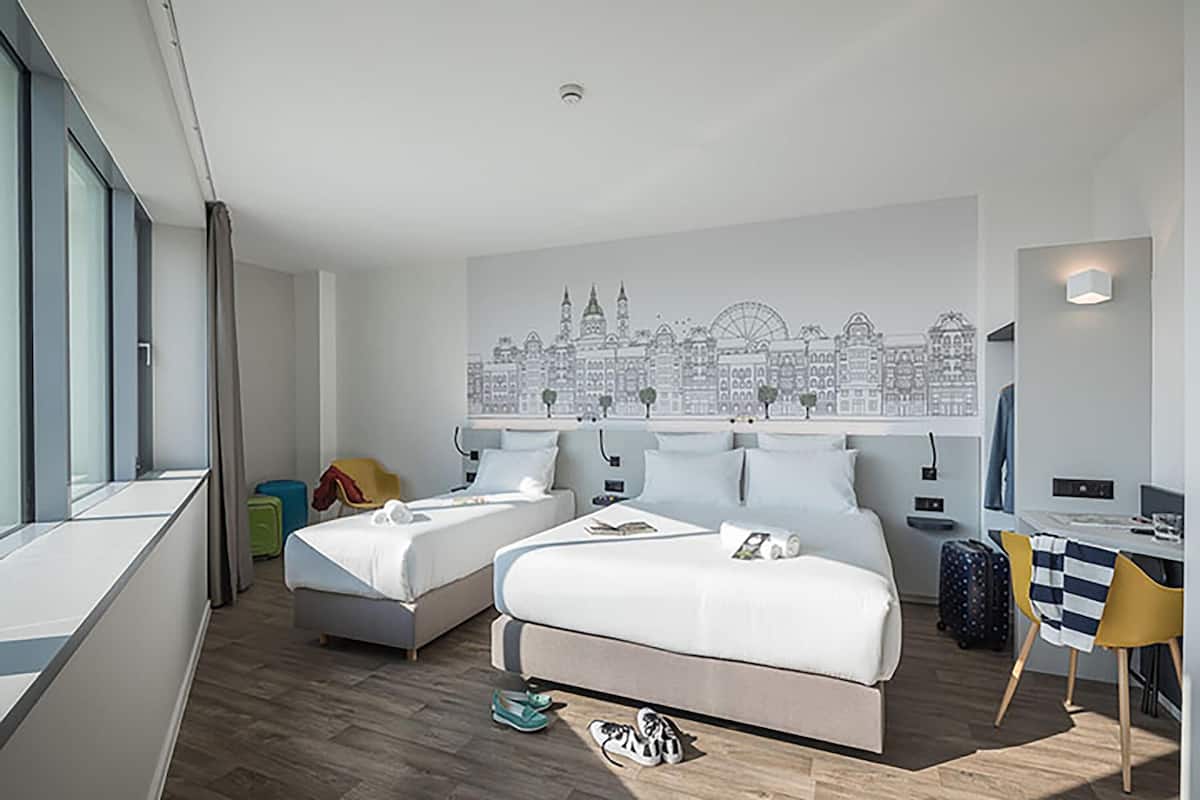 A modern hotel room with two beds, a cityscape mural, a desk, and a view of the city.