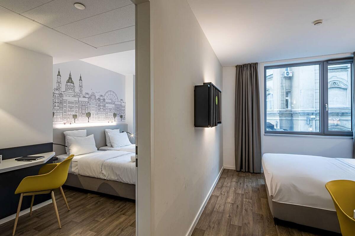 A hotel room with a bed, a desk, a chair, a TV, and a window with a view of a building.