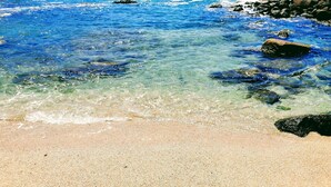Une plage avec une eau d’un bleu limpide, un rivage rocheux et un bâtiment visible au loin.