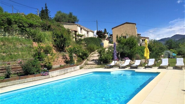 A rectangular swimming pool with clear blue water, surrounded by a tiled deck. There are several lounge chairs with yellow and purple umbrellas. The pool is set against a backdrop of a hillside with vegetation and buildings. - Studio (Bendejun)